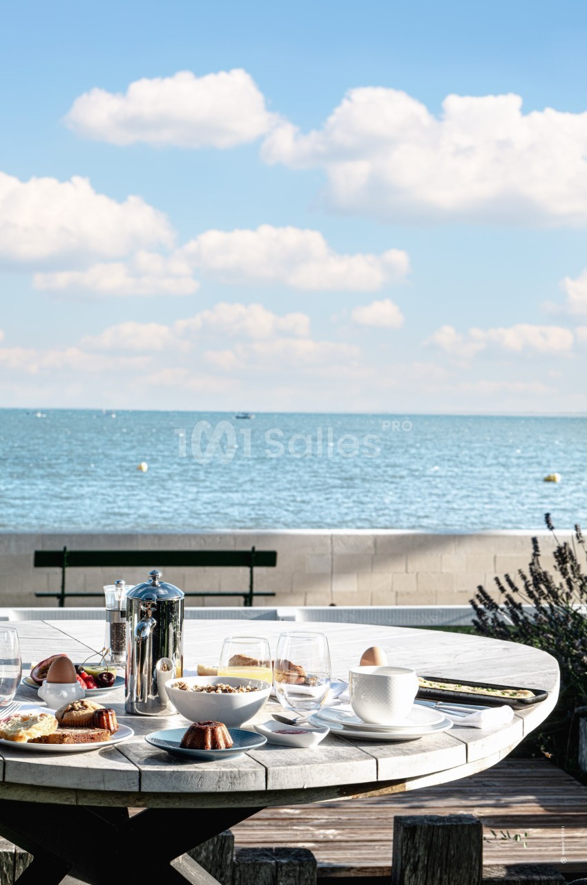 Table dressée pour un petit-déjeuner en extérieur avec vue sur la mer, sous un ciel bleu parsemé de nuages.