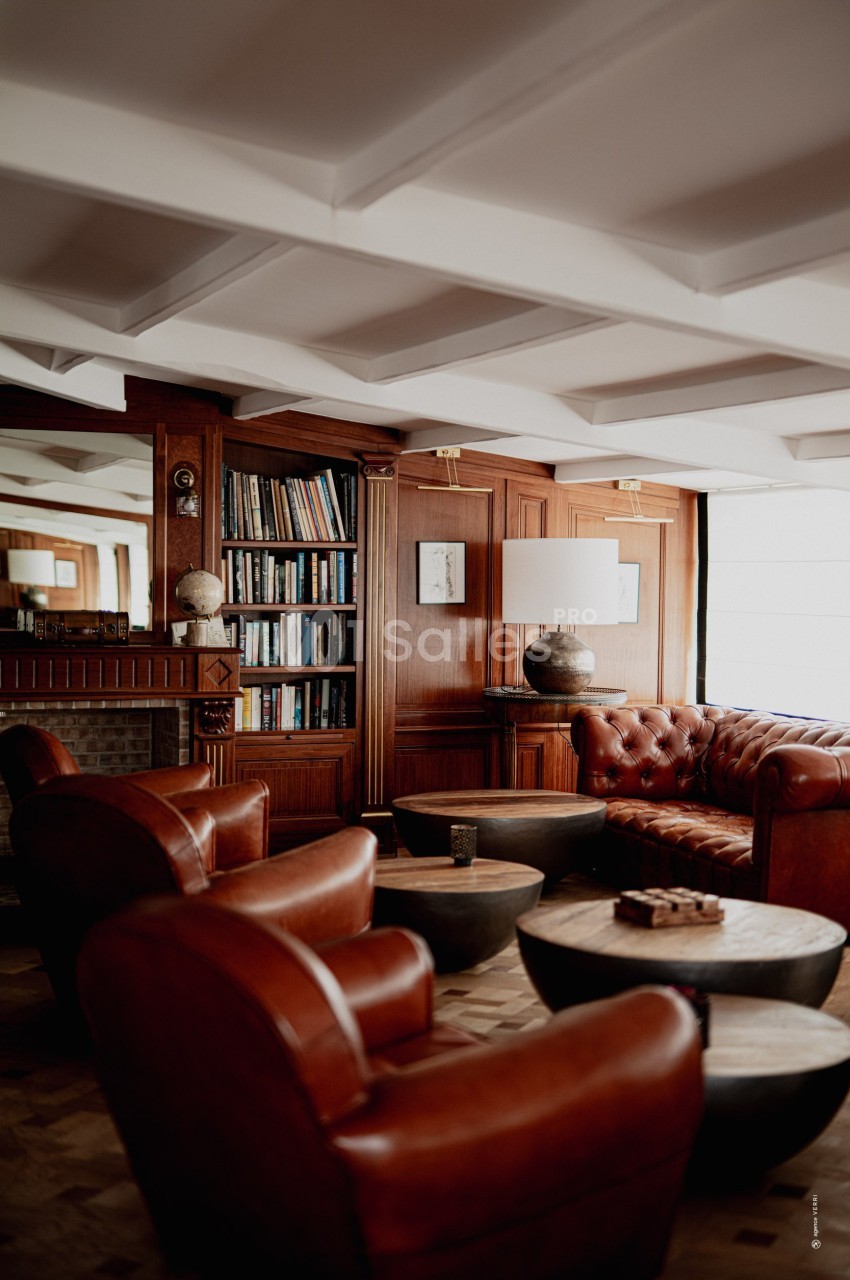Salon chaleureux avec fauteuils en cuir marron, tables basses en bois, bibliothèque et éclairage naturel.