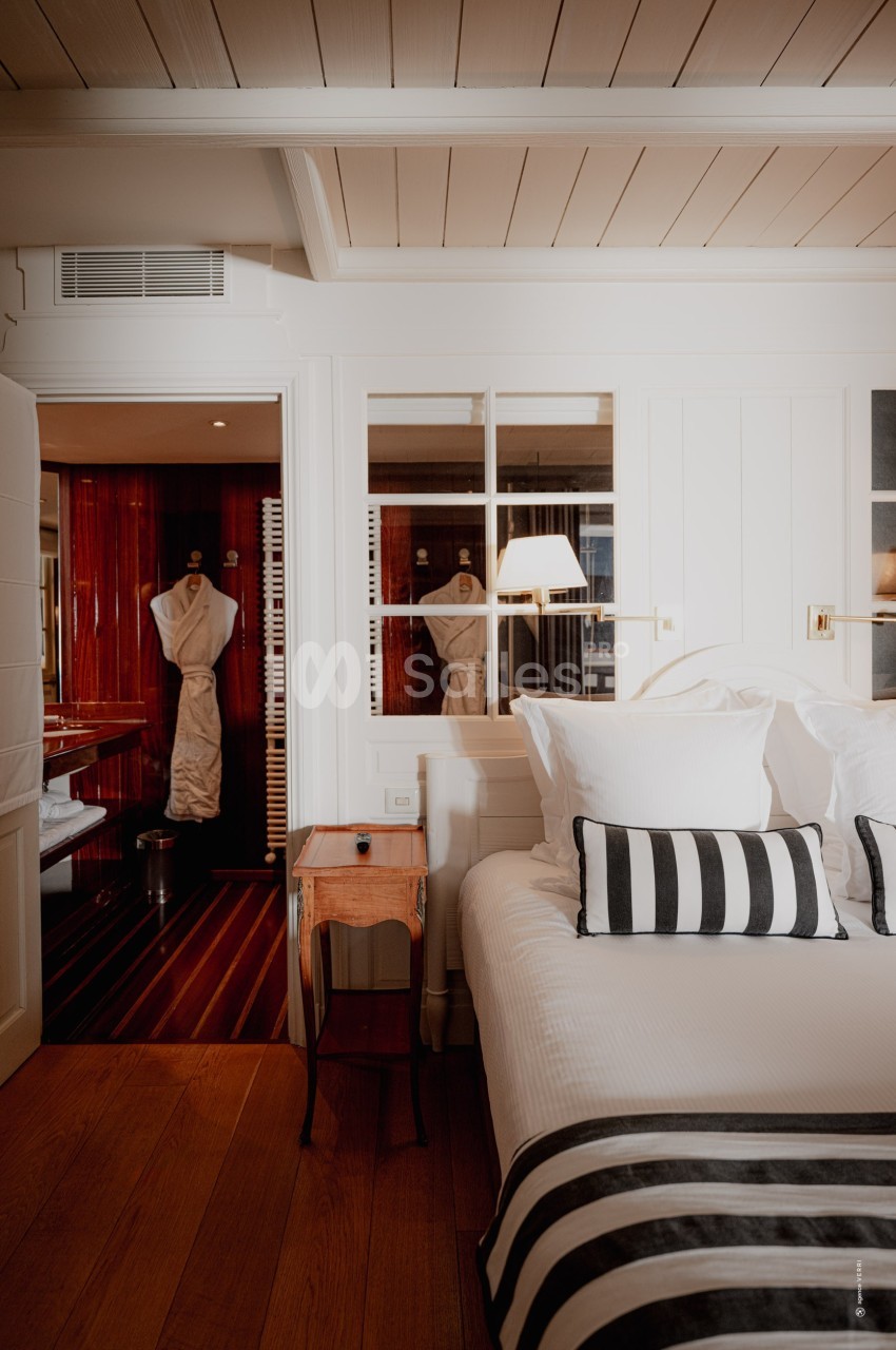 Chambre lumineuse avec lit double, oreillers et plaid rayés, vue sur une salle de bain en bois avec peignoir suspendu.