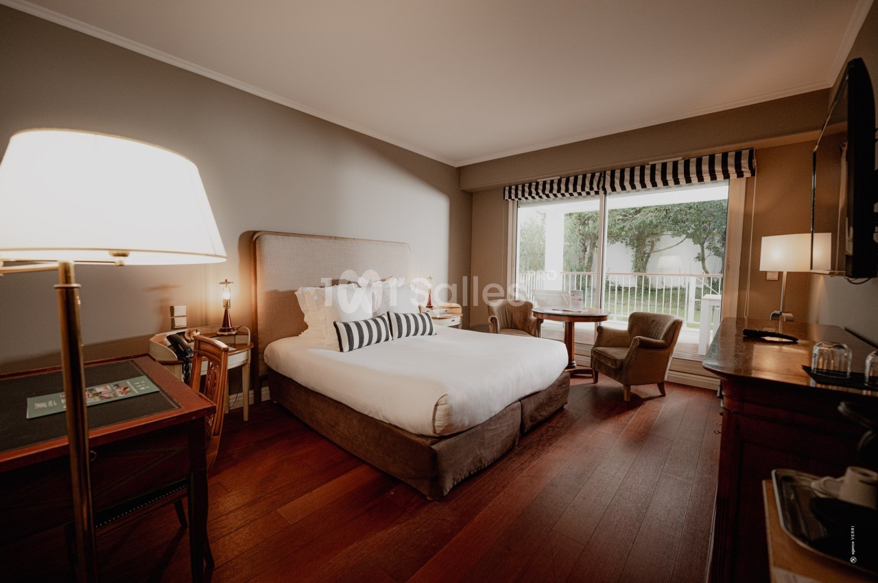 Chambre lumineuse avec lit double, bureau, fauteuils et grande baie vitrée donnant sur un jardin.