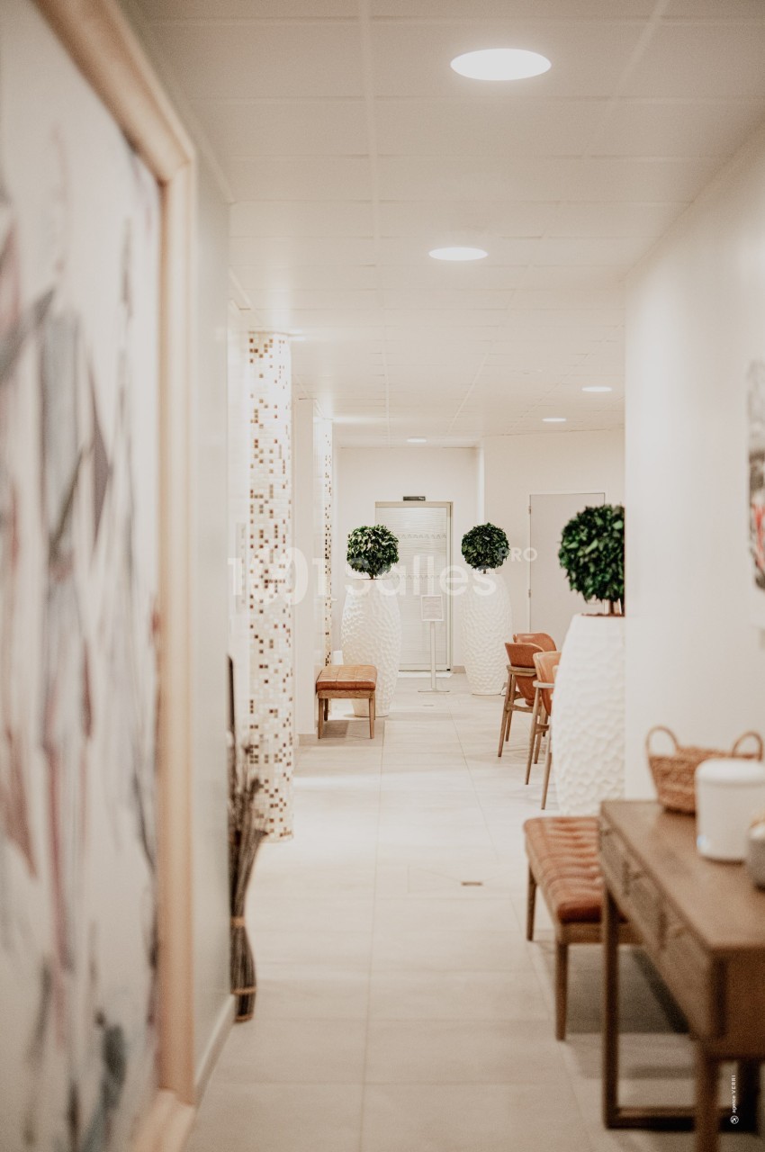 Couloir lumineux avec murs clairs, plantes décoratives en pots blancs, bancs en bois et carrelage beige.