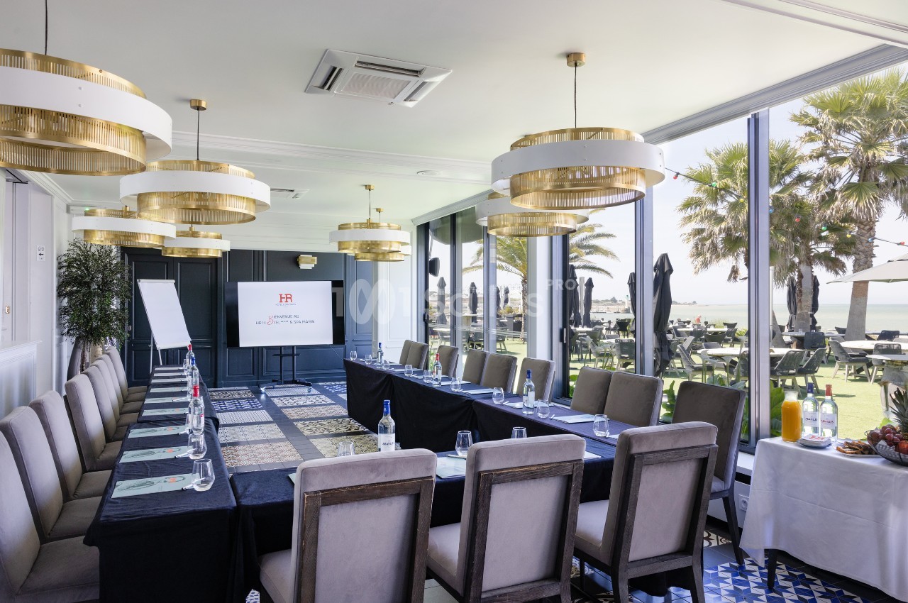 Salle de réunion lumineuse avec une grande table, chaises, écran de présentation et vue sur une terrasse et des palmiers.
