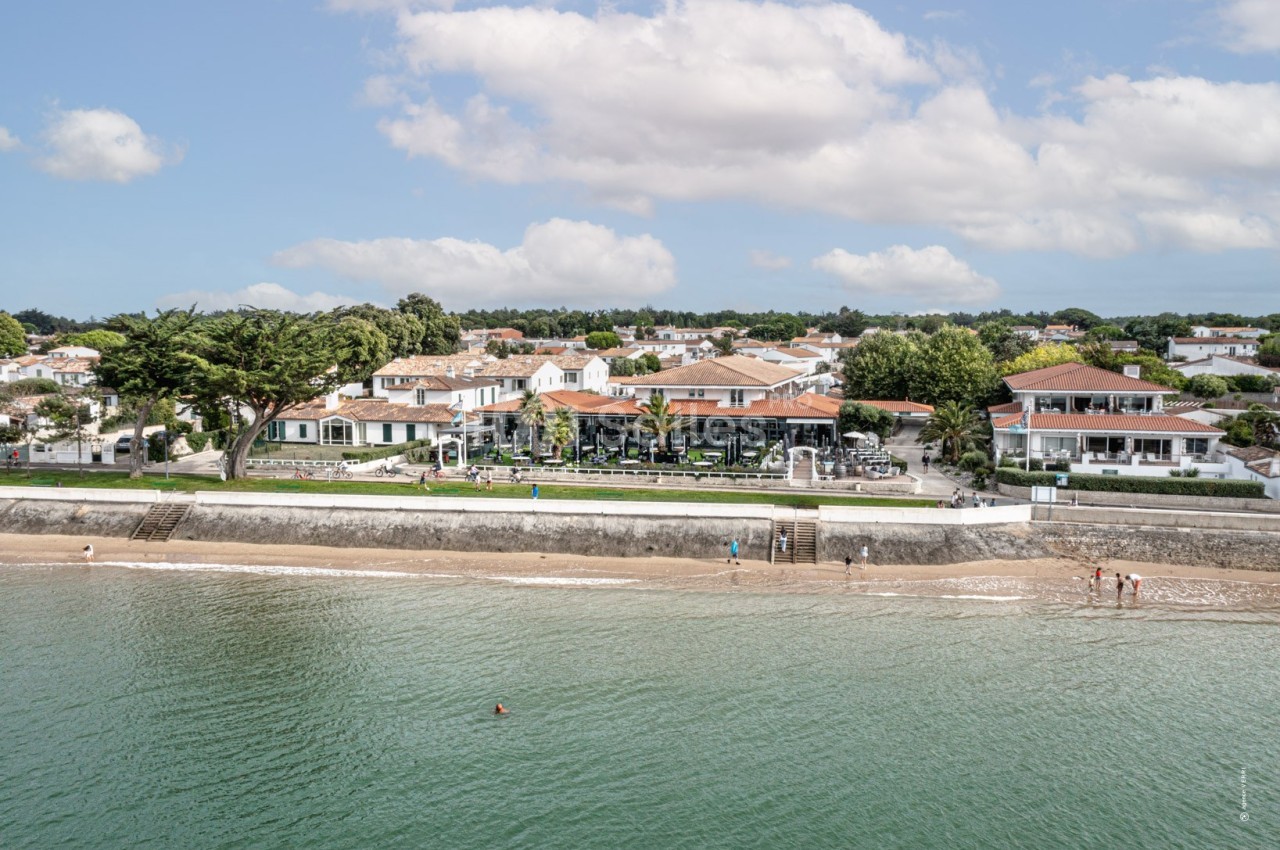 Vue aérienne d'une plage bordée de maisons blanches et végétation, avec des baigneurs au bord de l'eau.