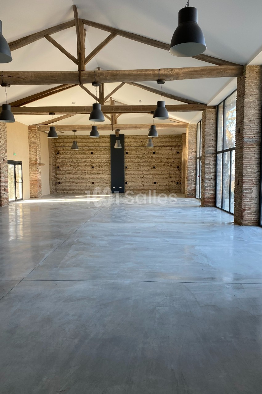 Salle spacieuse avec murs en briques, plafond à poutres apparentes et grandes baies vitrées laissant entrer la lumière…