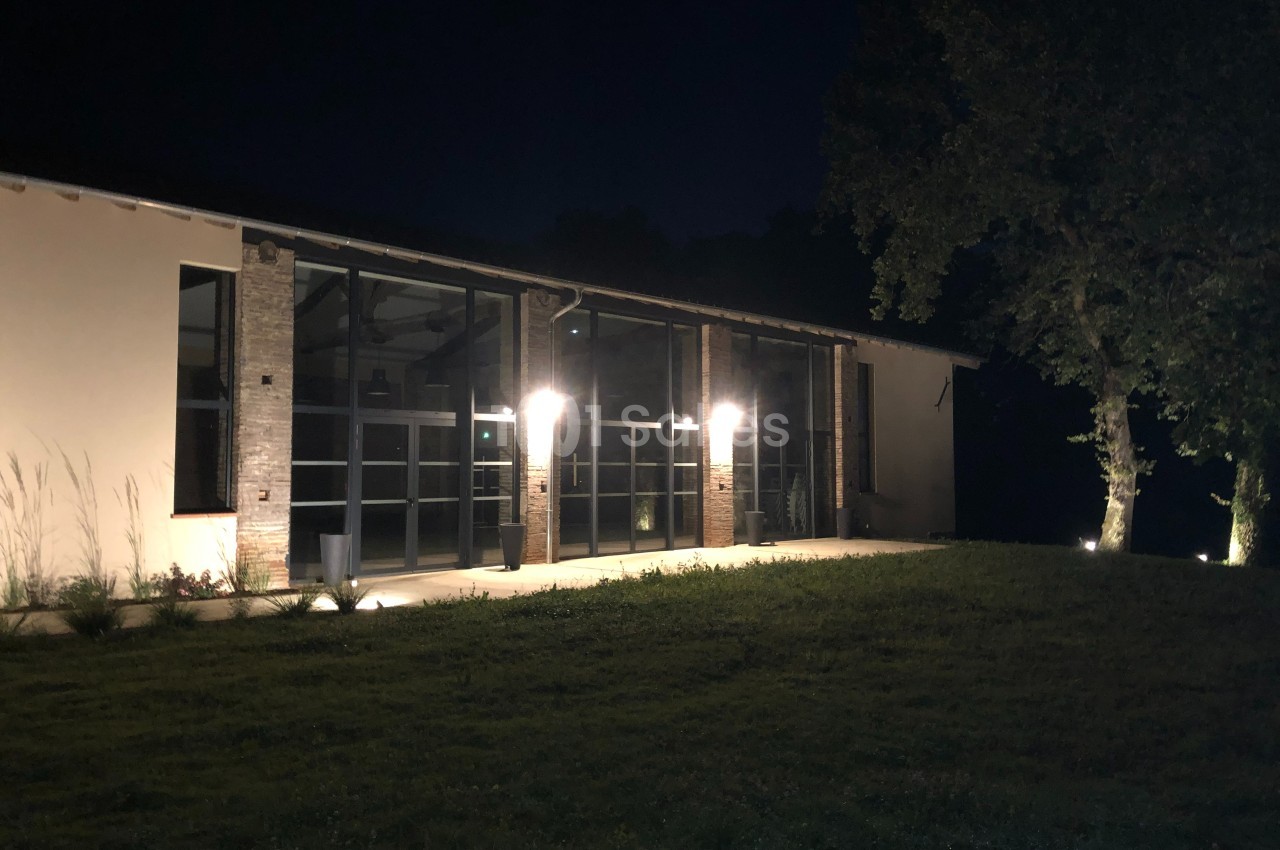 Façade d'un bâtiment moderne éclairé la nuit, avec des arbres et une pelouse au premier plan.