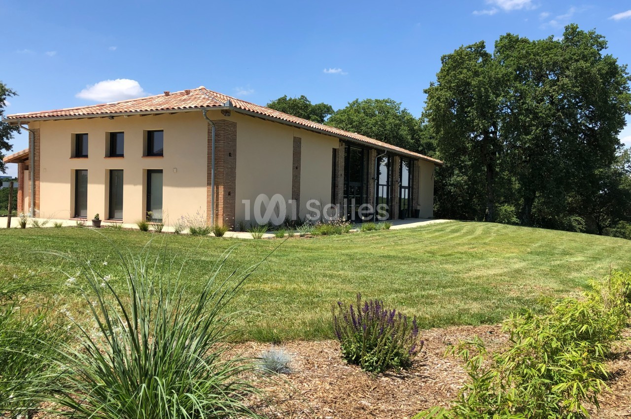 Maison moderne à un étage avec toit en tuiles, entourée de pelouse, plantes et arbres sous un ciel bleu.
