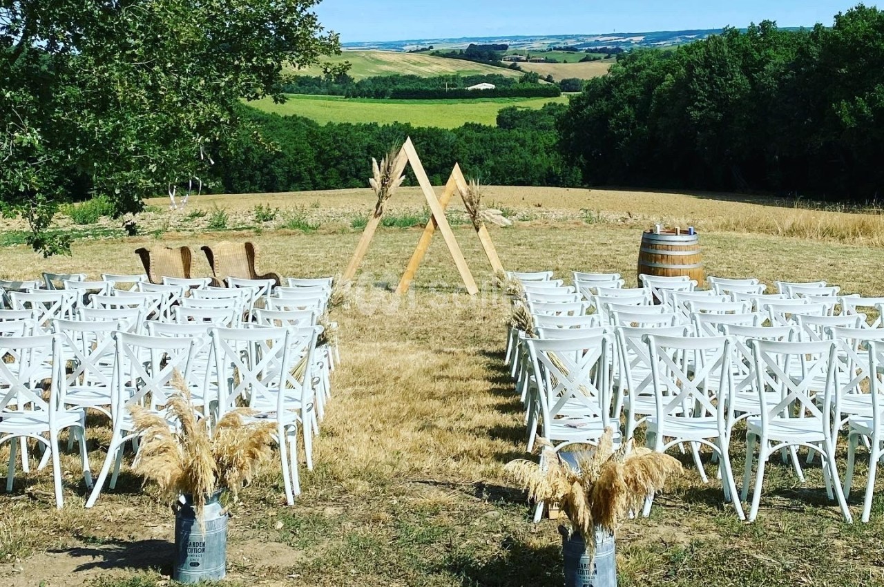 Chaises blanches alignées en extérieur face à une arche en bois triangulaire, entourées de champs et d'arbres.