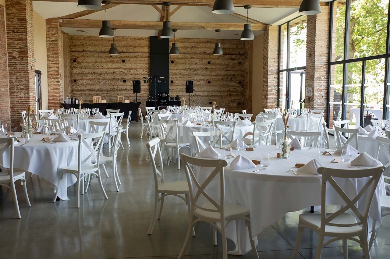 Salle de réception lumineuse avec tables rondes dressées de nappes blanches, chaises en bois et grandes baies vitrées.
