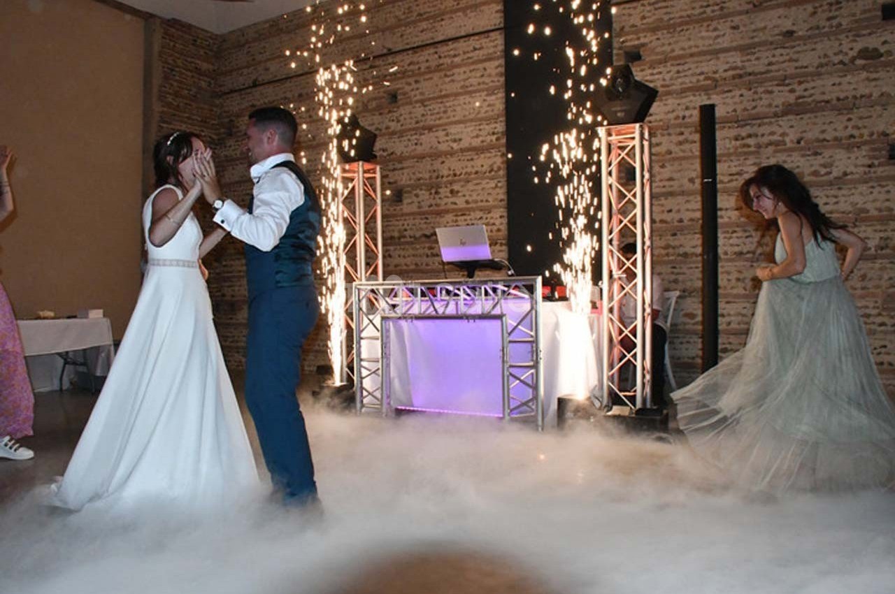 Un couple danse au milieu d'une piste entourée de fumée et d'effets pyrotechniques, avec une DJ en arrière-plan.