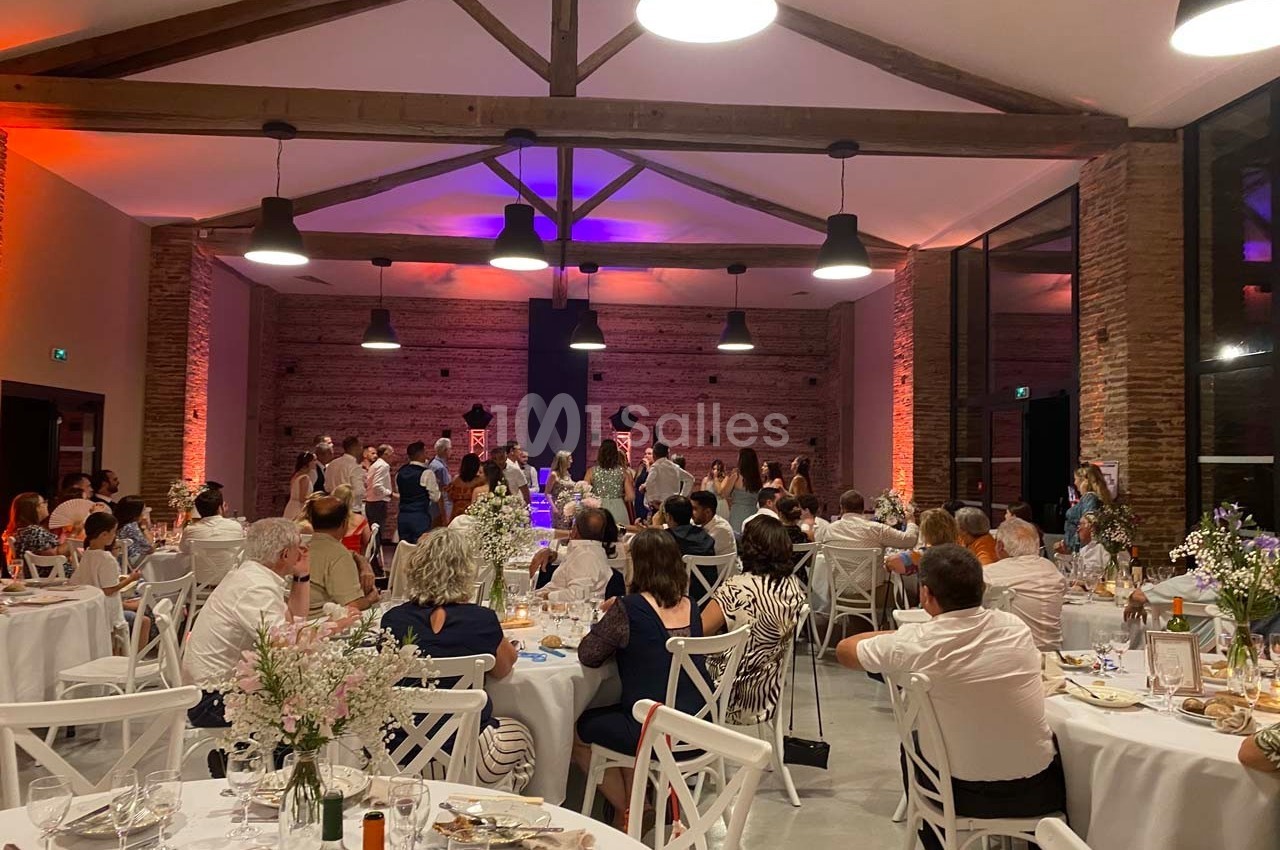 Salle de réception avec des invités assis et debout, éclairée par des lumières colorées et décorée de tables dressées.