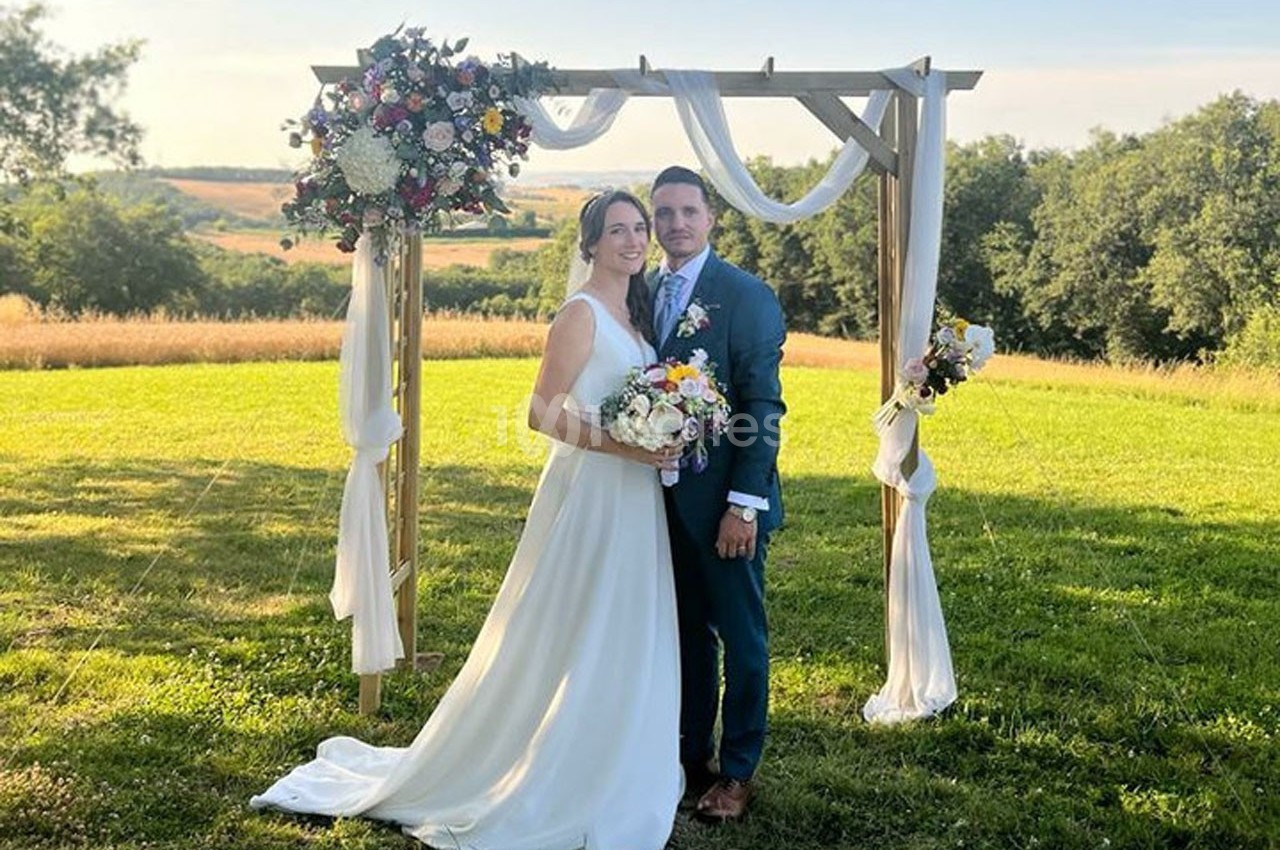Un couple en tenue de mariage pose sous une arche décorée de fleurs dans un champ verdoyant.