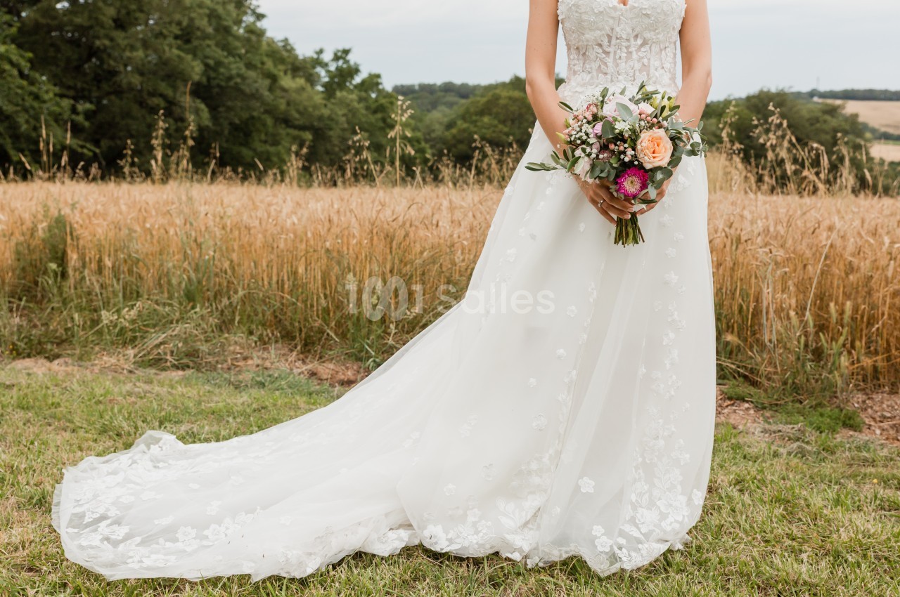 Robe de mariée blanche avec traîne, tenue par une femme tenant un bouquet coloré dans un champ en arrière-plan.
