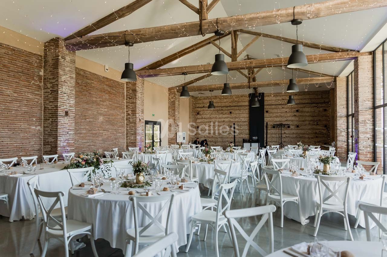 Salle de réception lumineuse avec murs en briques, poutres apparentes et tables rondes décorées pour un événement.