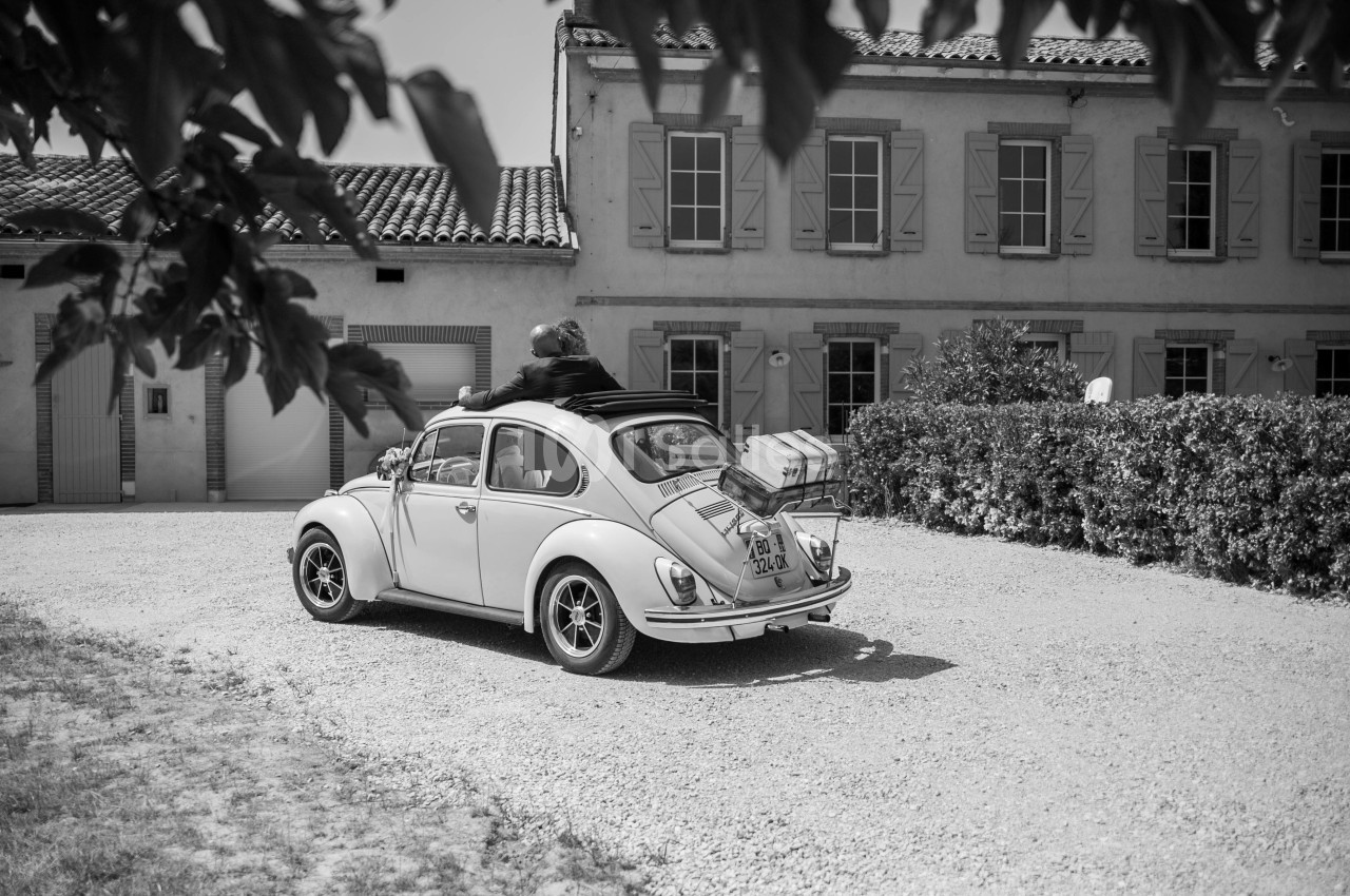 Voiture ancienne avec des bagages à l'arrière, garée devant une maison de campagne sous un ciel clair.