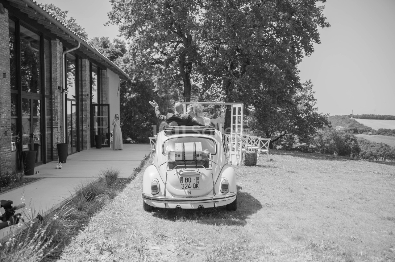 Un couple dans une voiture ancienne quitte un bâtiment entouré de verdure par une journée ensoleillée.