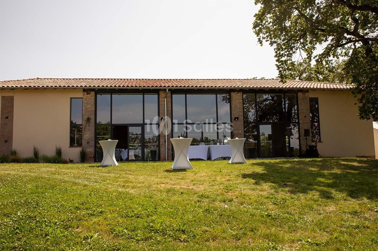 Façade d'un bâtiment moderne avec de grandes baies vitrées, entouré d'herbe et d'arbres, tables hautes à l'extérieur.