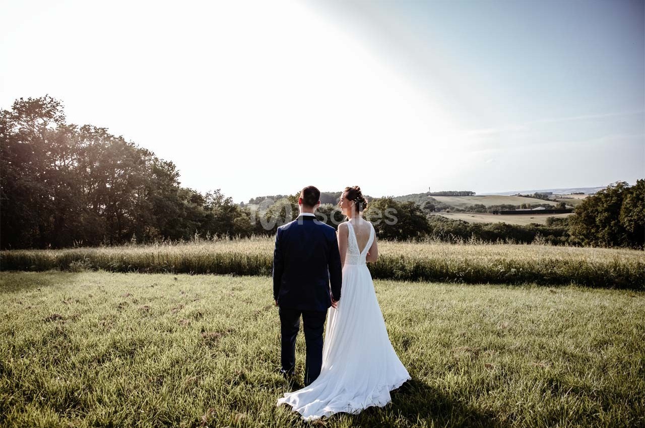 Un couple en tenue de mariage se tient debout dans un champ verdoyant, regardant un paysage vallonné.