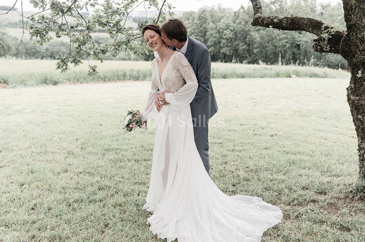 Un couple en tenue de mariage se tient sous un arbre dans un champ verdoyant, l'homme embrassant la femme.