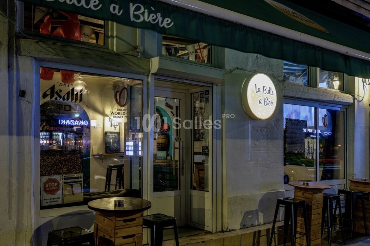 Façade d'un bar éclairé de nuit avec des tables hautes en bois disposées sur le trottoir.