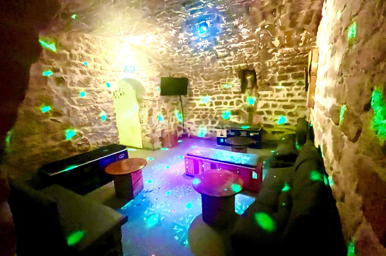 Salle voûtée en pierre éclairée par des lumières colorées, avec canapés, tables basses et écran mural.