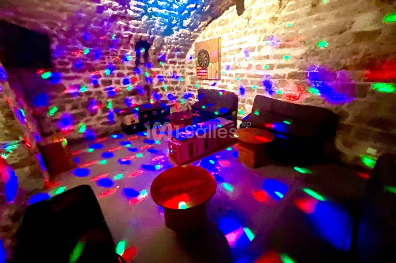 Salle voûtée en pierre éclairée par des lumières colorées, avec canapés, tables basses et cible de fléchettes au mur.