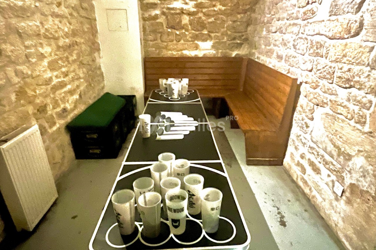 Table de beer-pong avec gobelets disposés, dans une pièce aux murs en pierre et bancs en bois.