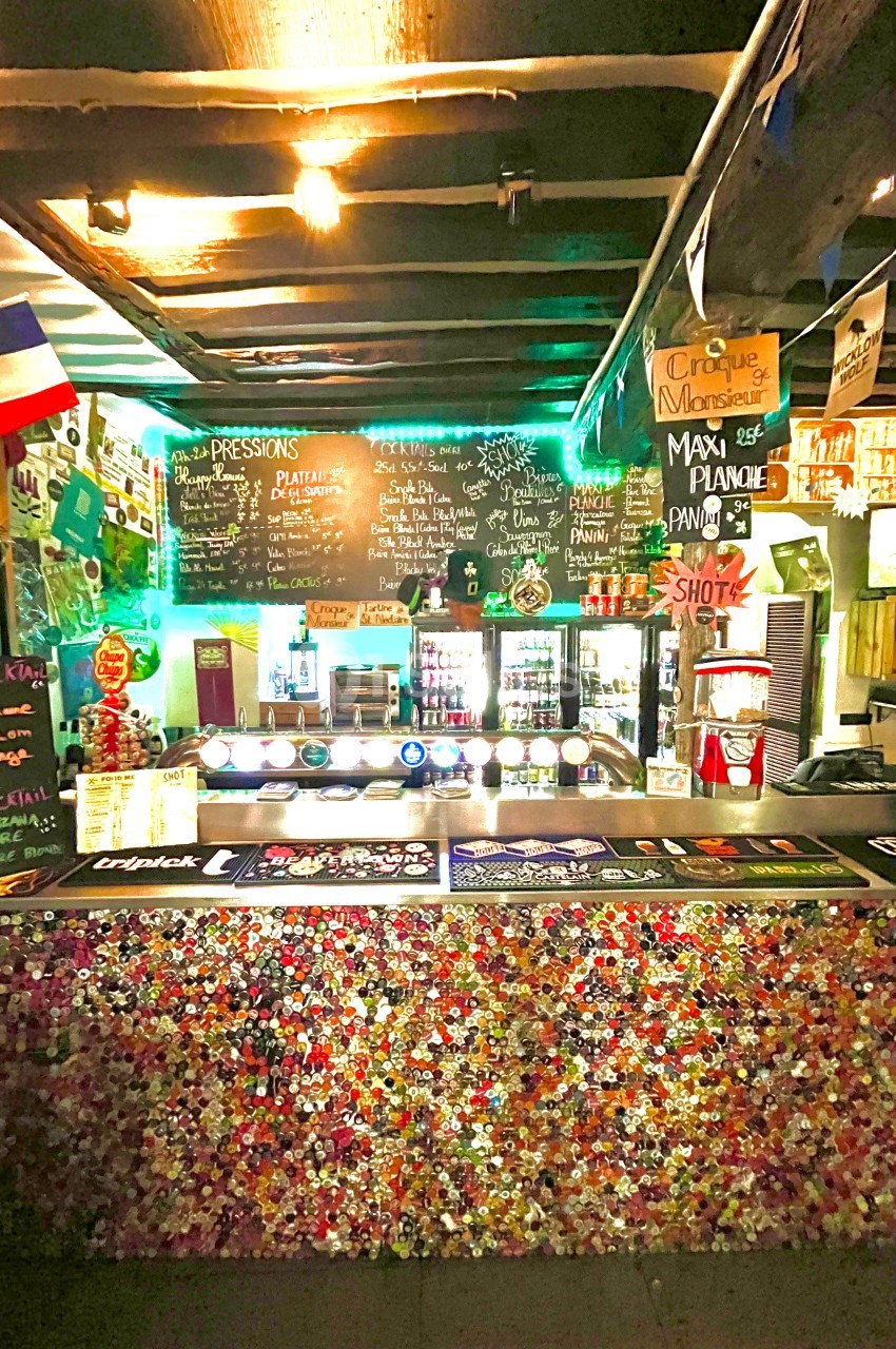 Comptoir de bar décoré de capsules de bouteilles, avec ardoises de menus, drapeaux et éclairage coloré en arrière-plan.