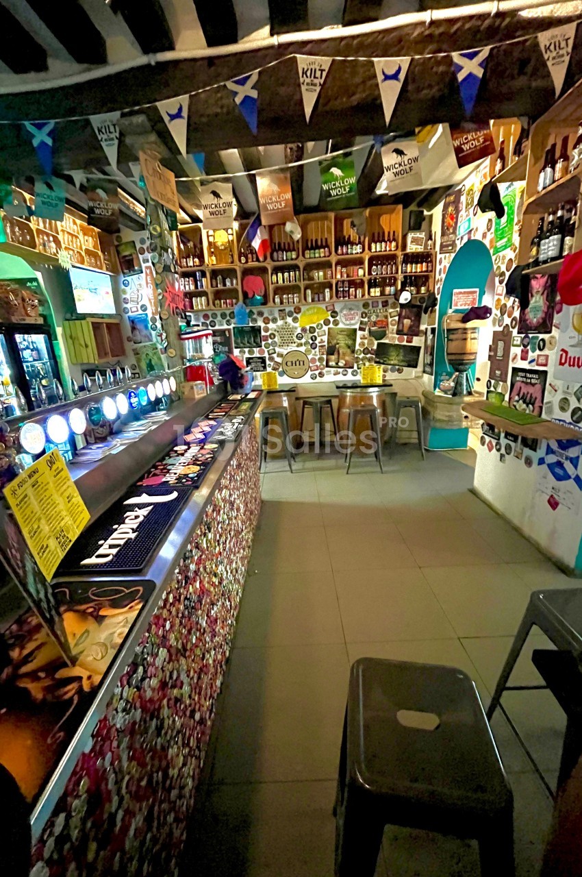 Intérieur d'un bar décoré avec des drapeaux, des bouteilles exposées et un long comptoir orné de capsules.