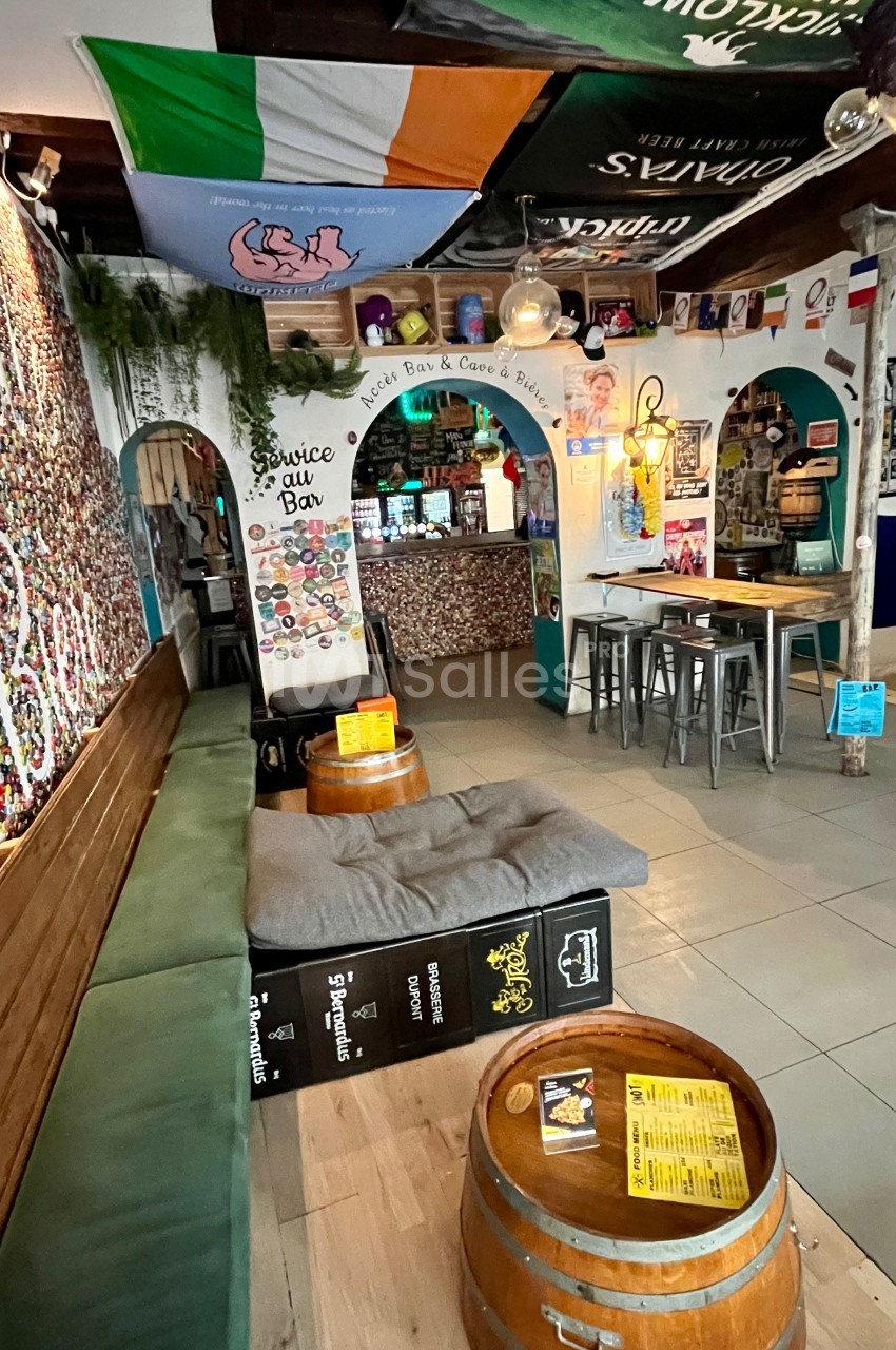 Intérieur d'un bar avec des tonneaux en tables, un banc, des drapeaux suspendus et un comptoir décoré au fond.