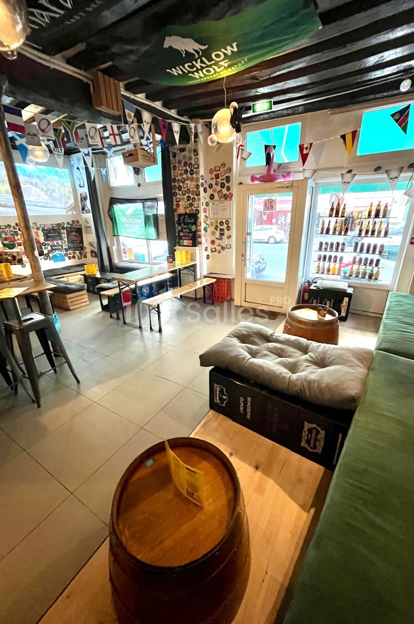 Intérieur d'un bar avec tables en bois, banquettes, décorations murales et étagères remplies de bouteilles.