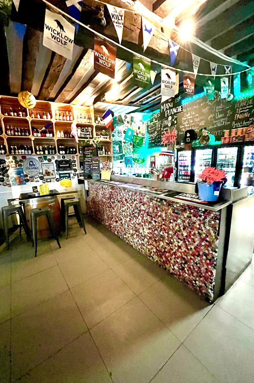Intérieur d'un bar avec comptoir décoré de capsules, étagères remplies de bouteilles et drapeaux suspendus au plafond.