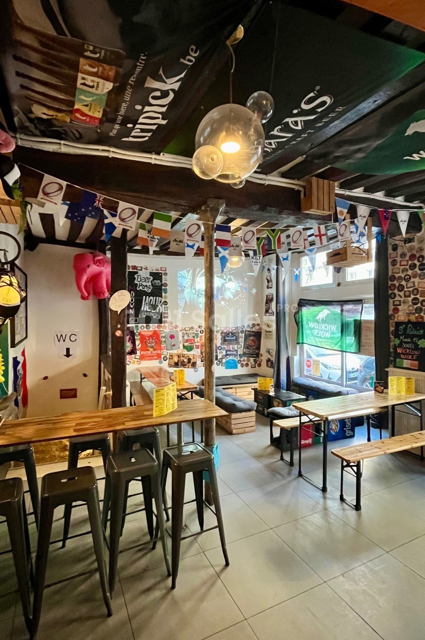 Intérieur d'un bar décoré de fanions, affiches et stickers, avec des tables en bois et des tabourets métalliques.