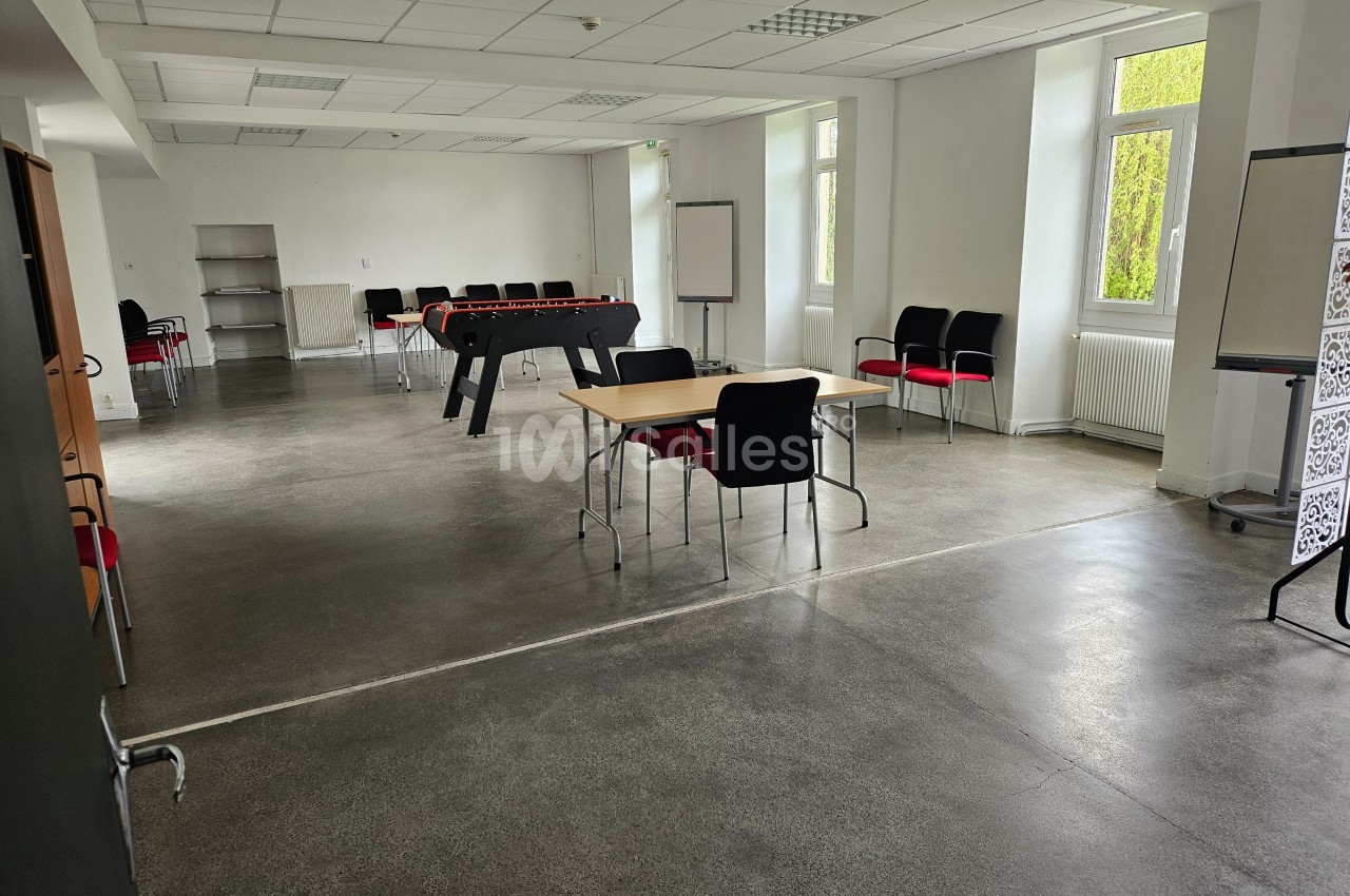 Salle lumineuse avec tables, chaises, baby-foot et tableaux blancs, sol en béton poli et grandes fenêtres.