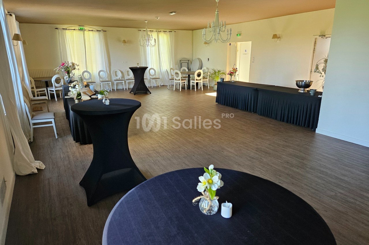 Salle lumineuse avec tables hautes noires, chaises blanches alignées et décoration florale sobre.