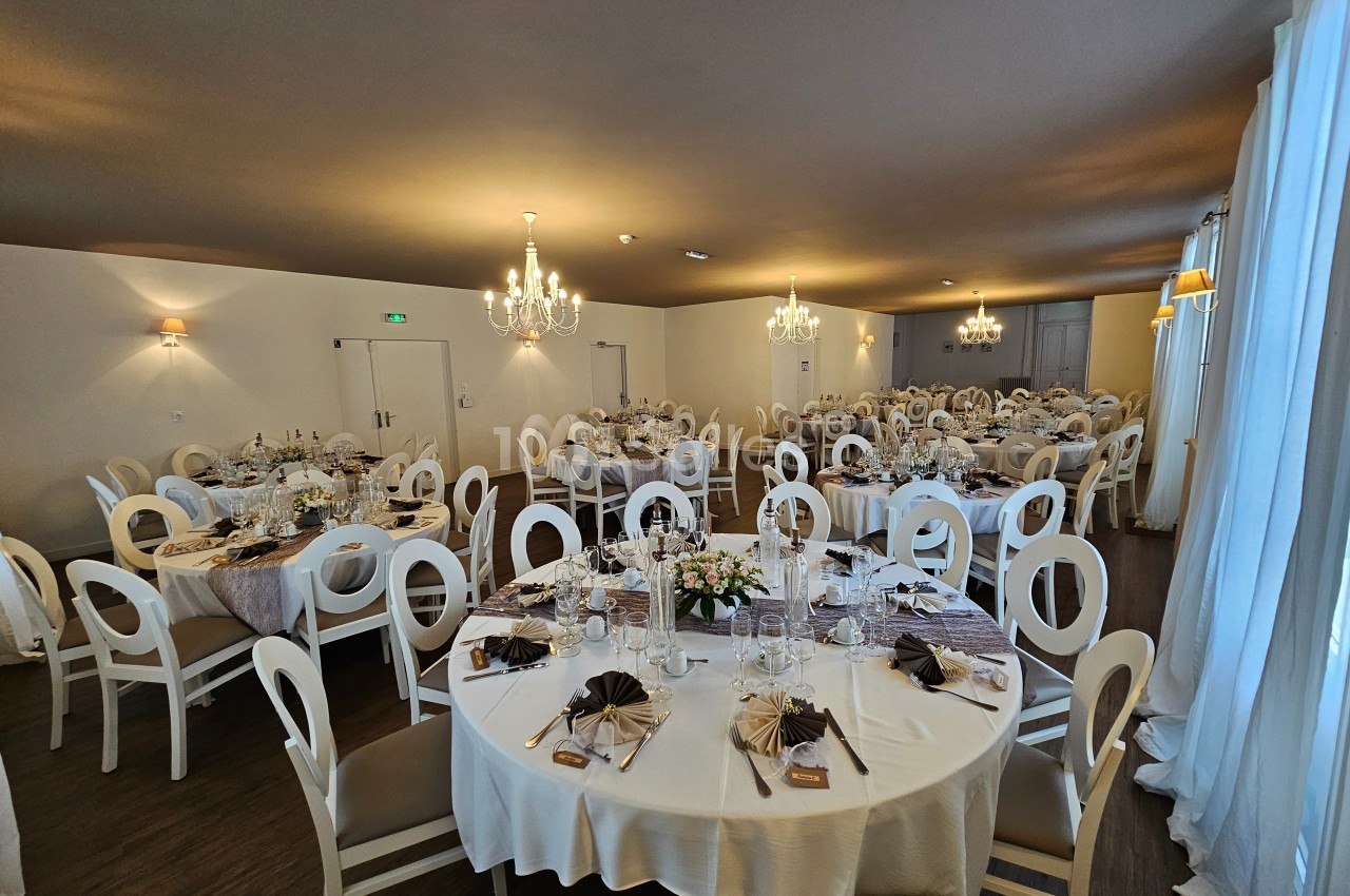 Salle de réception élégamment décorée avec des tables rondes dressées, chaises blanches et lumière naturelle.
