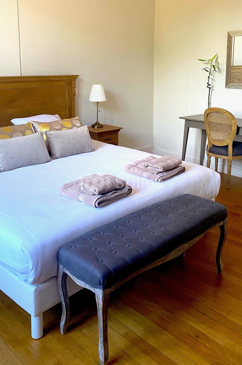 Chambre lumineuse avec lit double, banc capitonné, bureau en bois, lampe et miroir sur un parquet en bois clair.