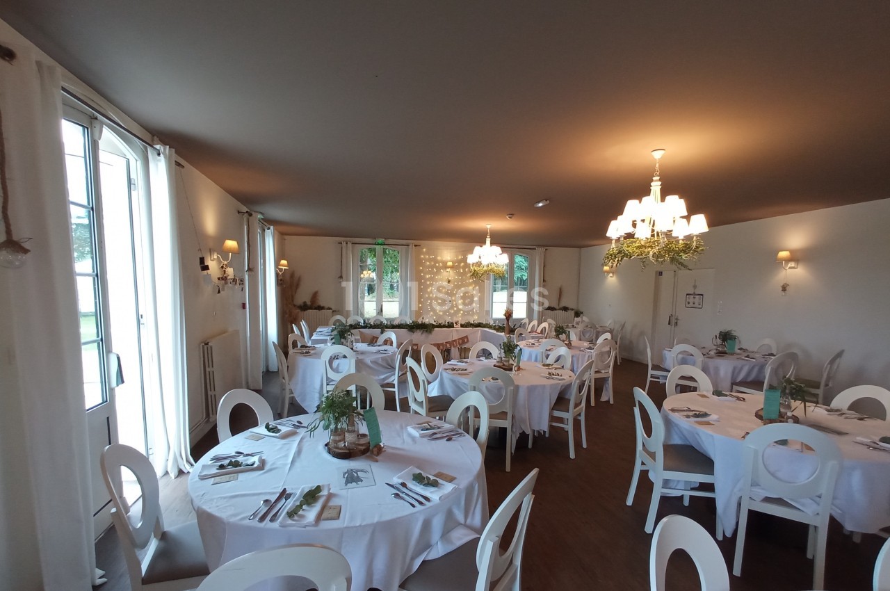 Salle de réception décorée avec des tables rondes dressées, chaises blanches et éclairage chaleureux.