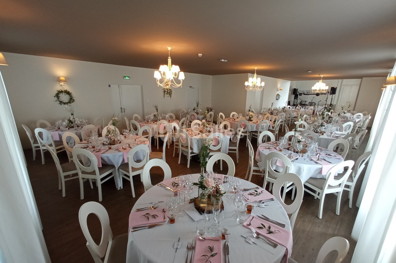 Salle de réception décorée avec des tables rondes, nappes blanches, serviettes roses et centres de table fleuris.