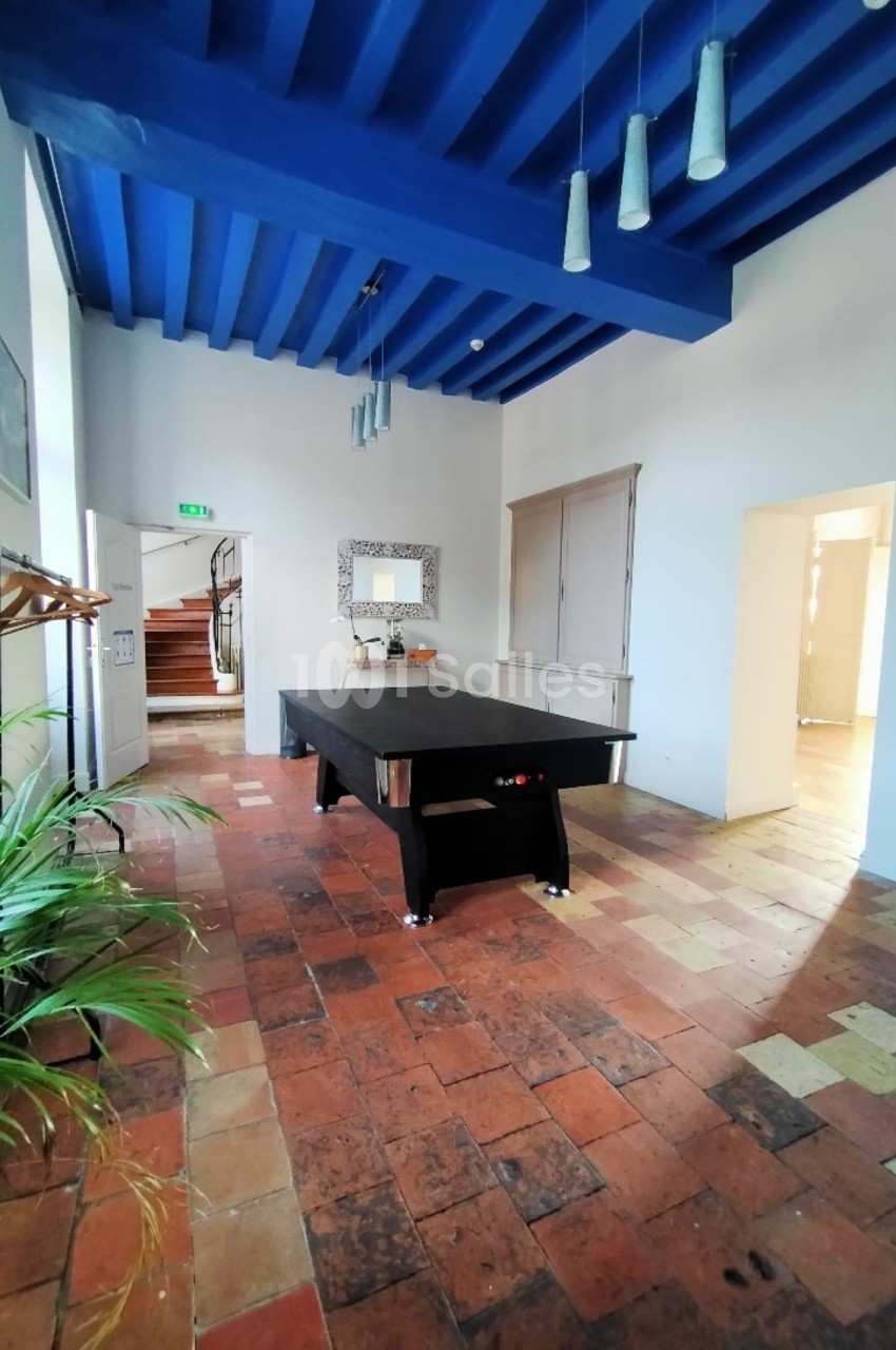 Salle lumineuse avec un billard noir au centre, plafond bleu, sol en carreaux anciens et plantes en décoration.