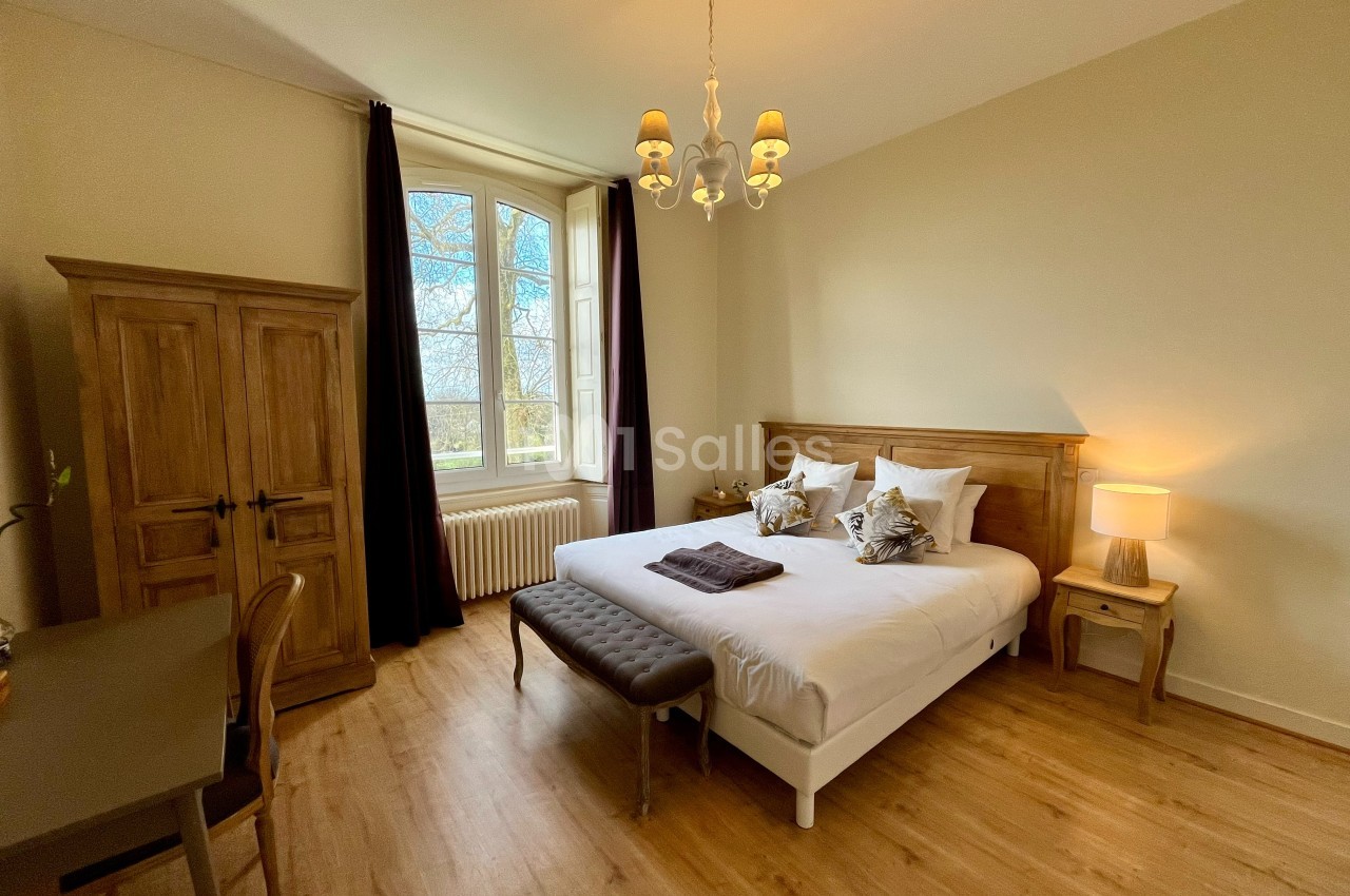 Chambre lumineuse avec lit double, armoire en bois, bureau, et fenêtre donnant sur un paysage arboré.