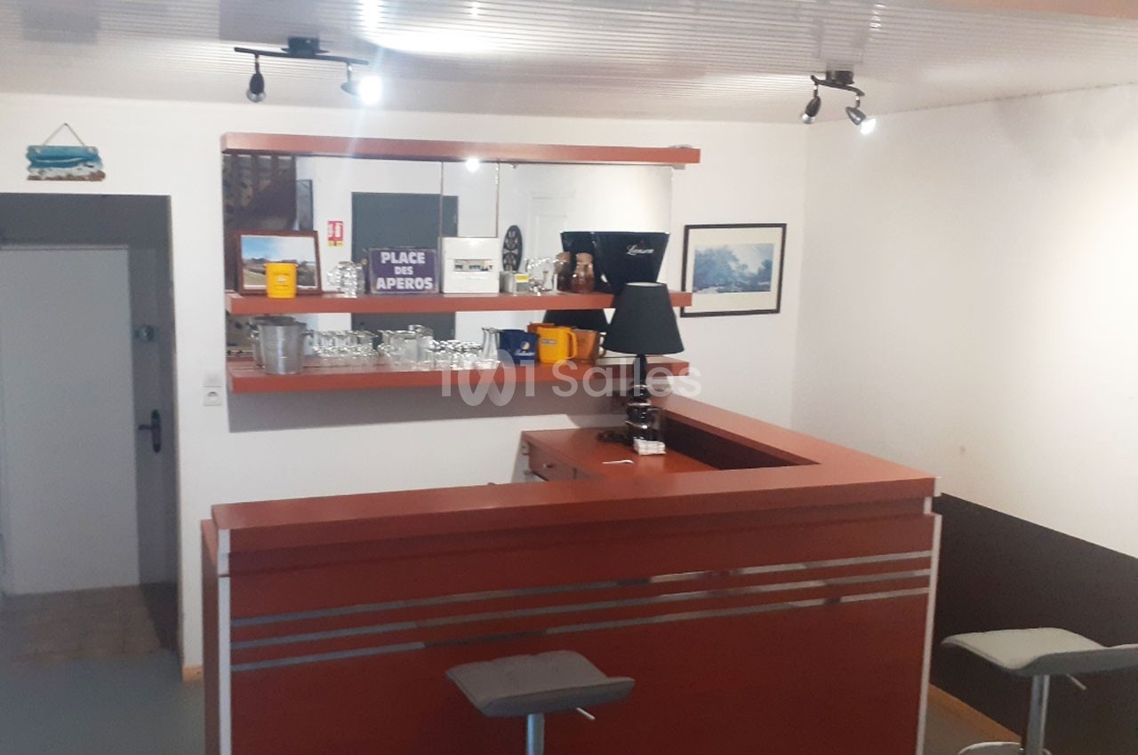 Comptoir de bar en bois avec tabourets, étagères contenant verres et objets décoratifs, éclairage au plafond.