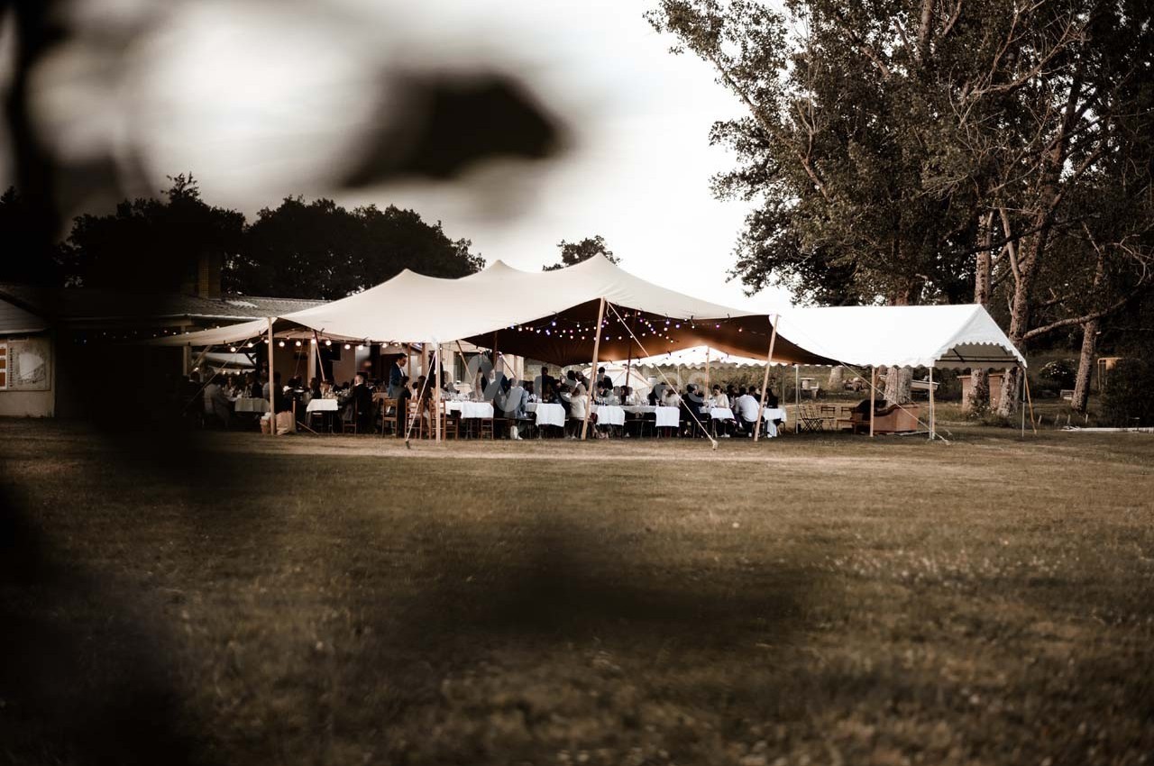 Tente blanche installée en extérieur avec des invités assis à des tables, entourée d'arbres et d'une pelouse.