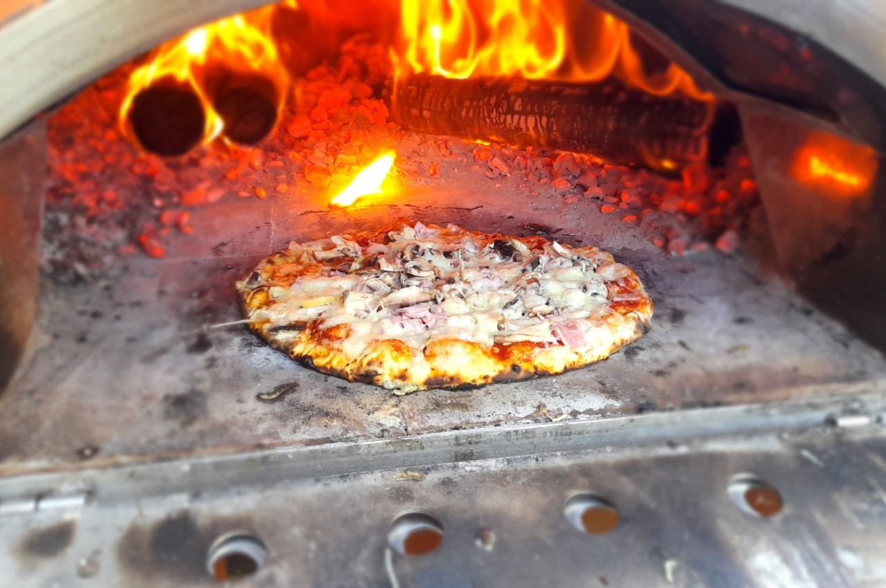 Pizza cuisant dans un four à bois avec des flammes vives et des braises incandescentes en arrière-plan.
