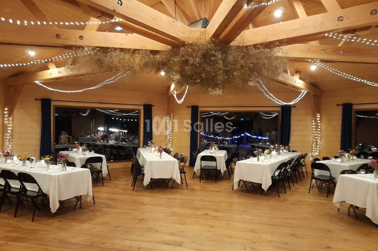 Salle en bois décorée pour un événement, avec tables dressées, guirlandes lumineuses et grande structure en poutres…