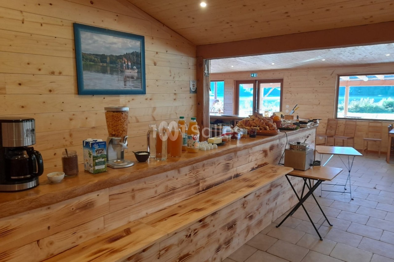 Buffet de petit-déjeuner avec boissons, céréales, viennoiseries et pain dans une salle en bois lumineuse.