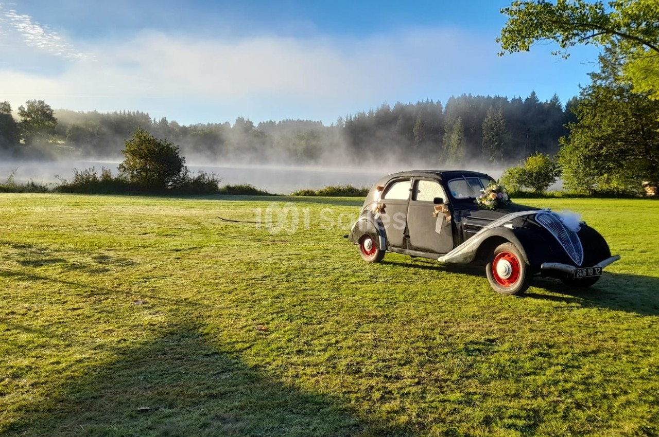 Voiture ancienne noire décorée pour un événement, stationnée sur une pelouse au bord d'un lac entouré de brume matinale.