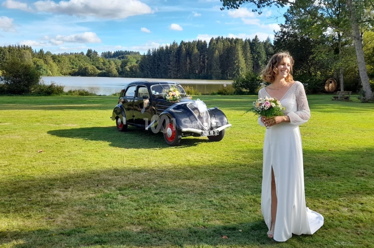 Une femme en robe de mariée tient un bouquet sur une pelouse, avec une voiture ancienne et un lac en arrière-plan.
