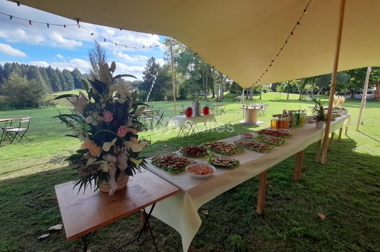 Buffet en plein air sous une tente, avec des plats variés, des boissons et une décoration florale sur une table.
