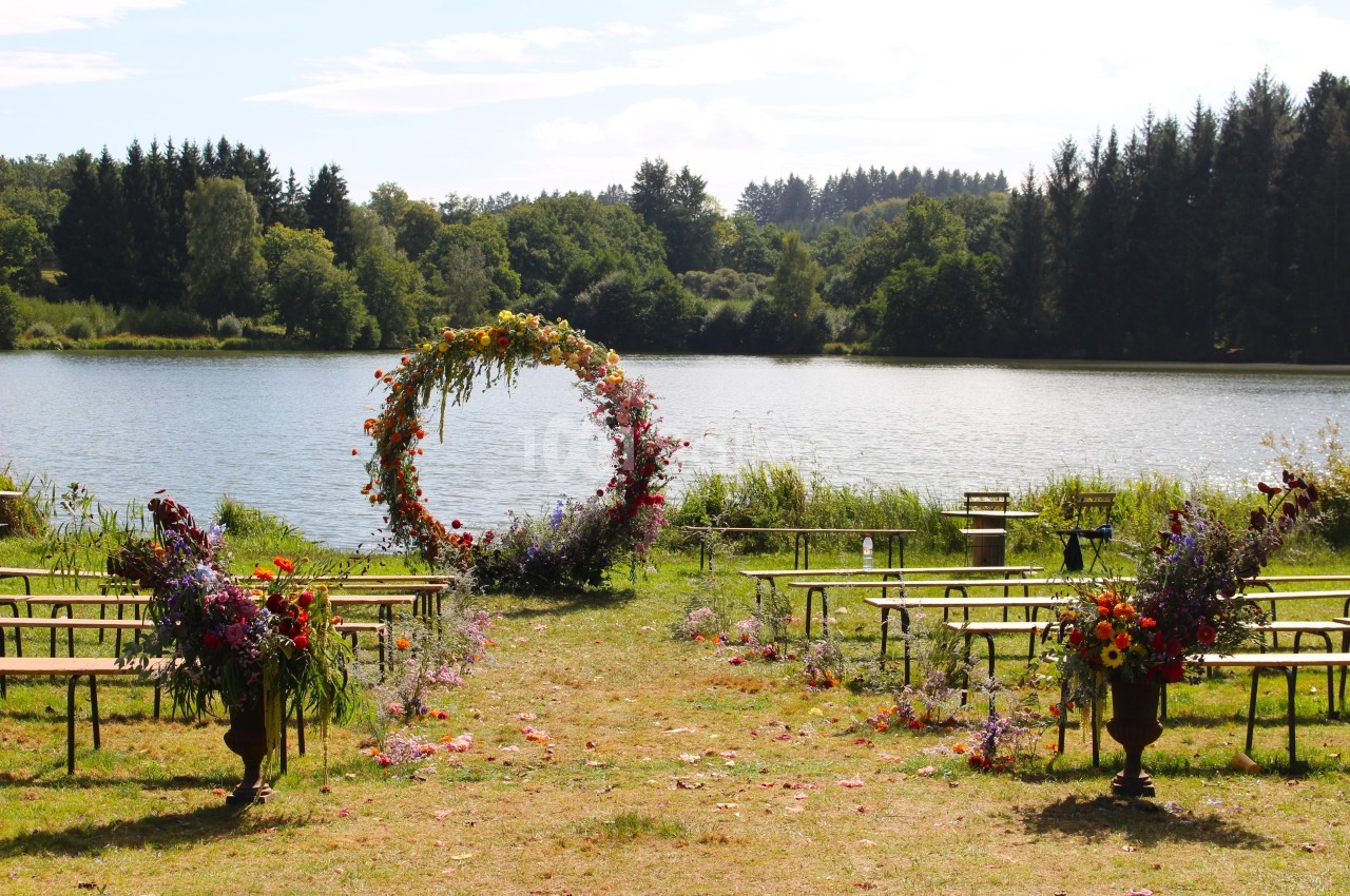 Cérémonie en plein air avec arche florale circulaire, bancs en bois et vue sur un lac entouré de forêt.