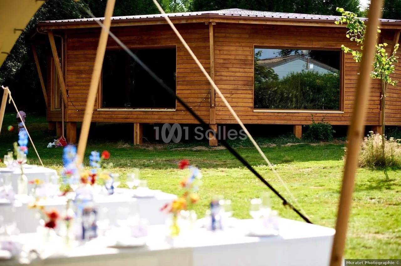 Salle en bois avec de grandes fenêtres, entourée de verdure, avec des tables décorées pour un événement au premier plan.